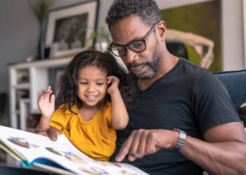 A loving father of African descent sits on the couch at home and reads a storybook to his preschool age daughter. The child is sitting on her father's lap and is smiling while looking at the book.