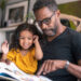 A loving father of African descent sits on the couch at home and reads a storybook to his preschool age daughter. The child is sitting on her father's lap and is smiling while looking at the book.