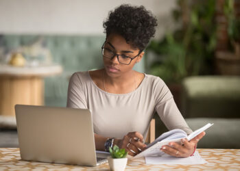 Focused young african american businesswoman or student looking at laptop holding book learning, serious black woman working or studying with computer doing research or preparing for exam online